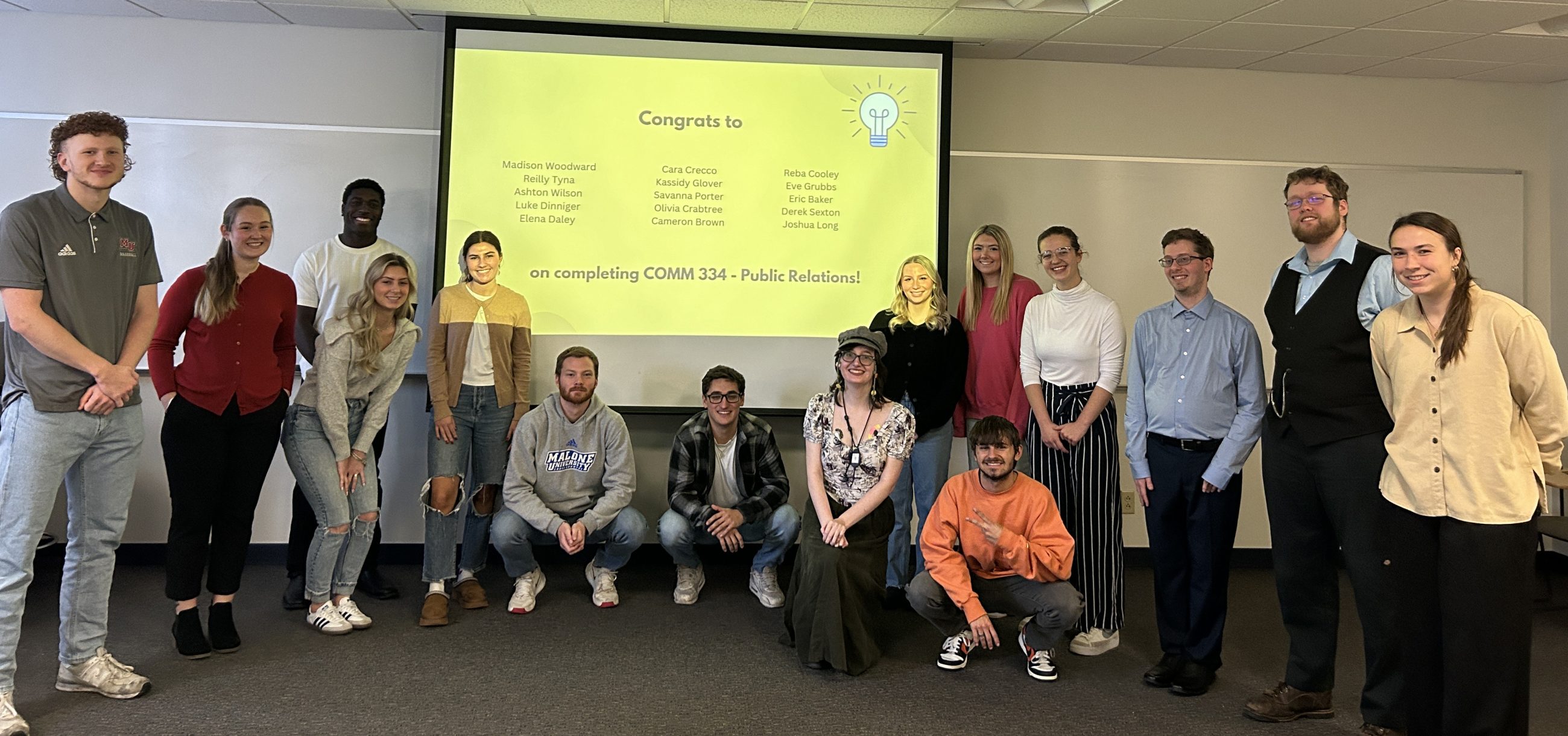 15 communication arts students gathered in a semicircle in front of a screen showing award recipients' names