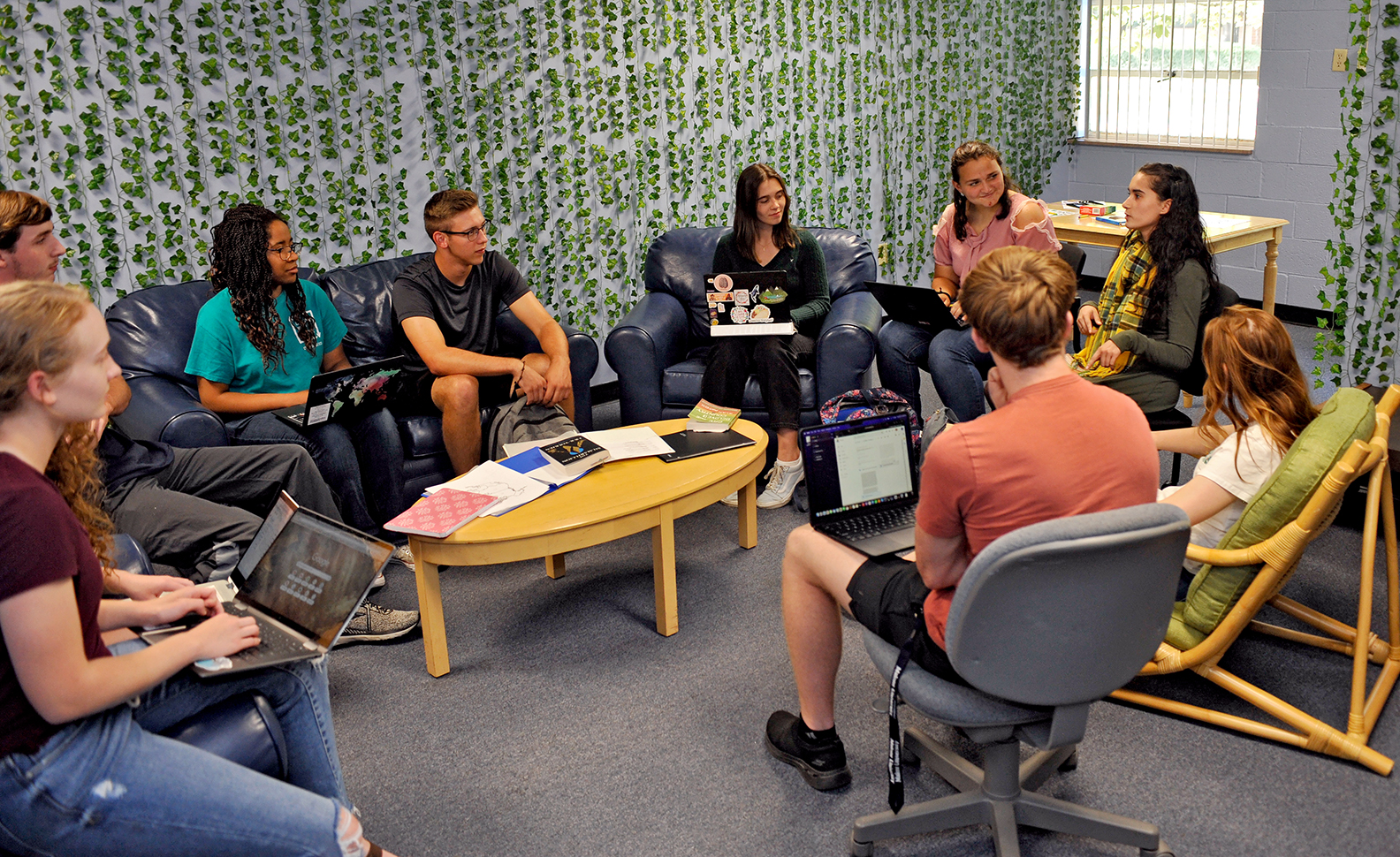 9 males and females seated in circle several of whom have open laptops; one appears to be speaking to the rest of the group