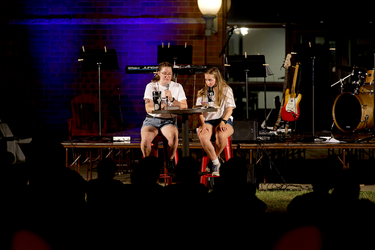 2 females in front of outdoor platform containing musical instruments; silhouettes of audience in foreground