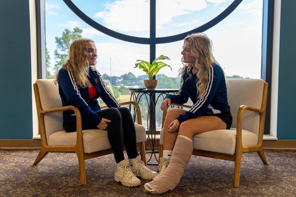 2 female students seated indoor chairs in front of large round window; 1 of the students with leg/foot in cast up to knee_3 (close-up view)