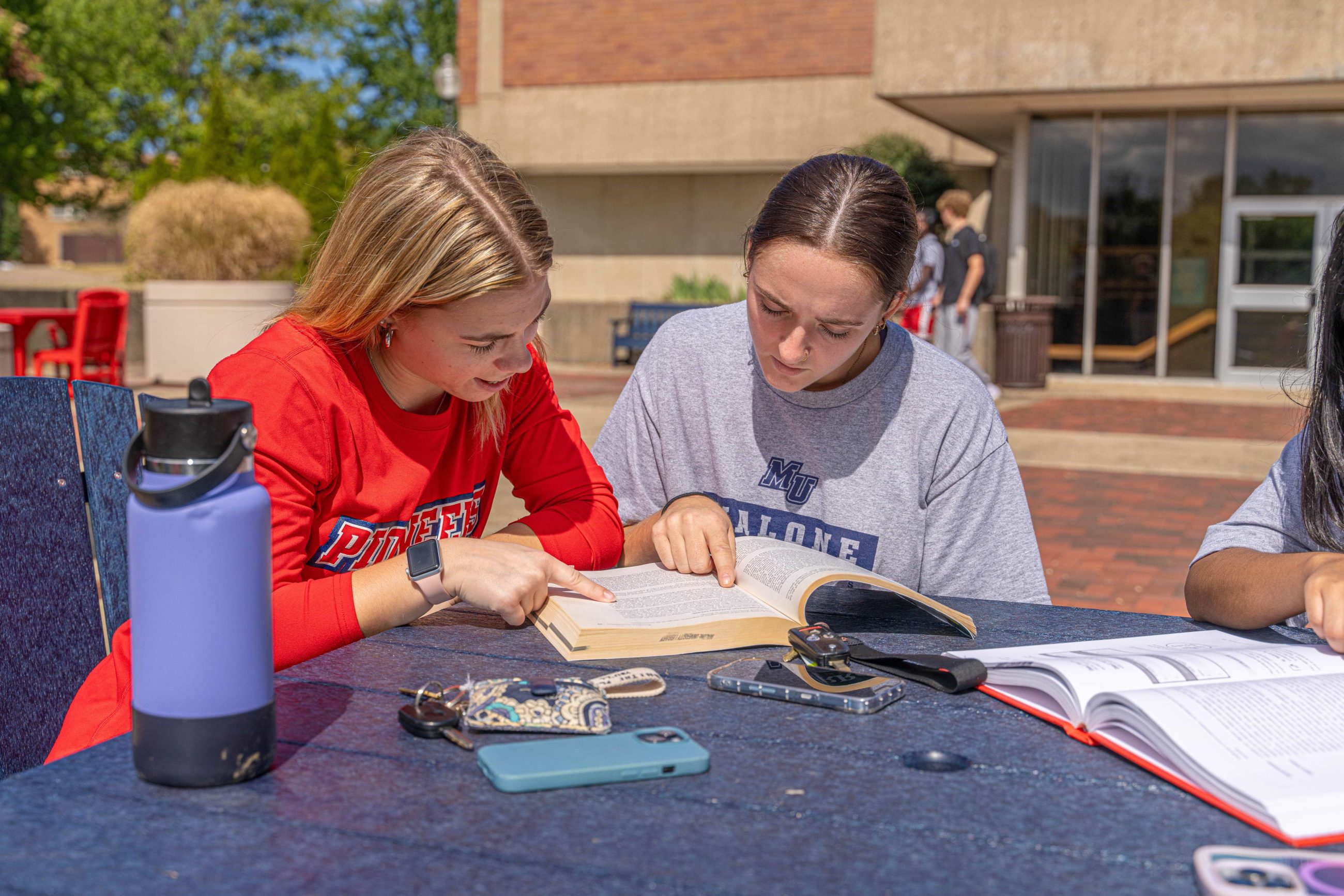 2 females studying a page from a book at table in front of Cattell Library