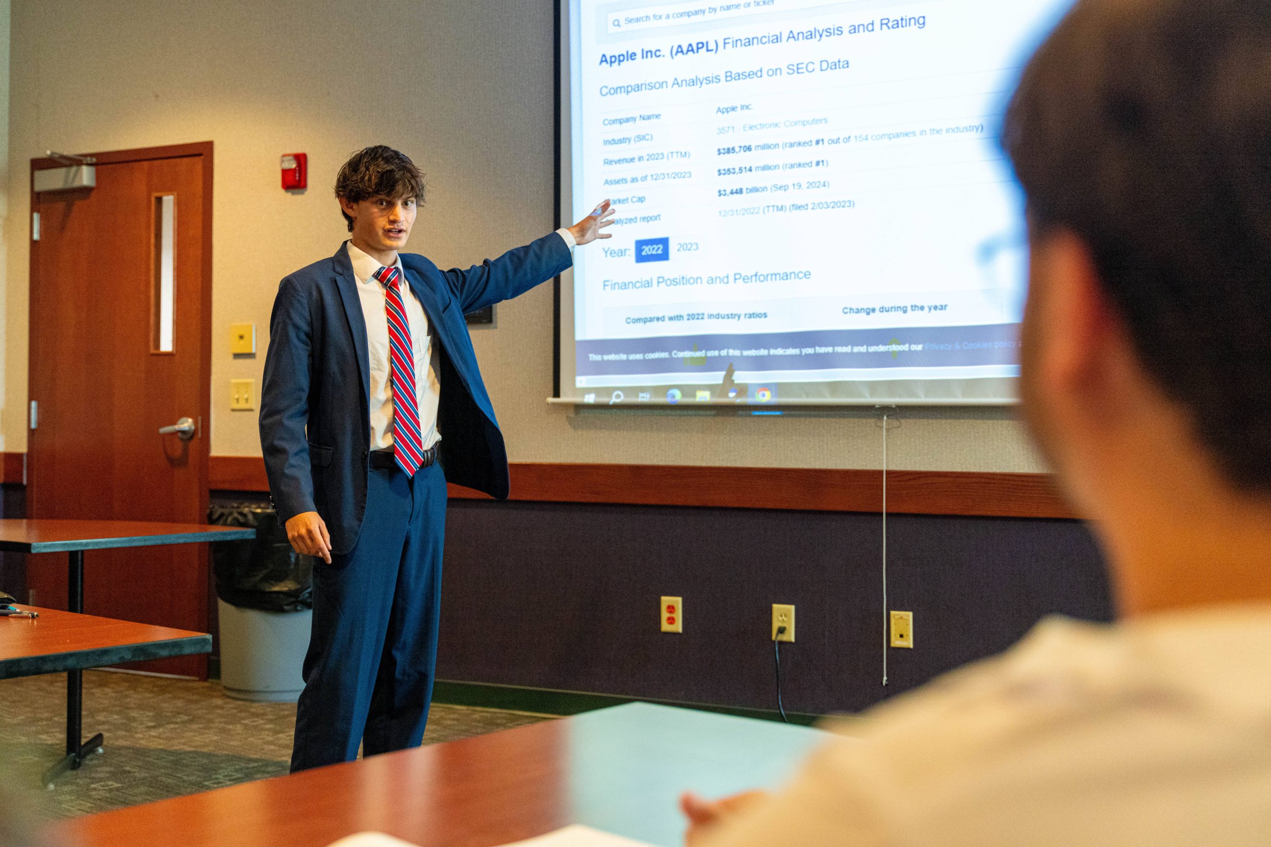 male student in suit gesturing toward image on a wall screen