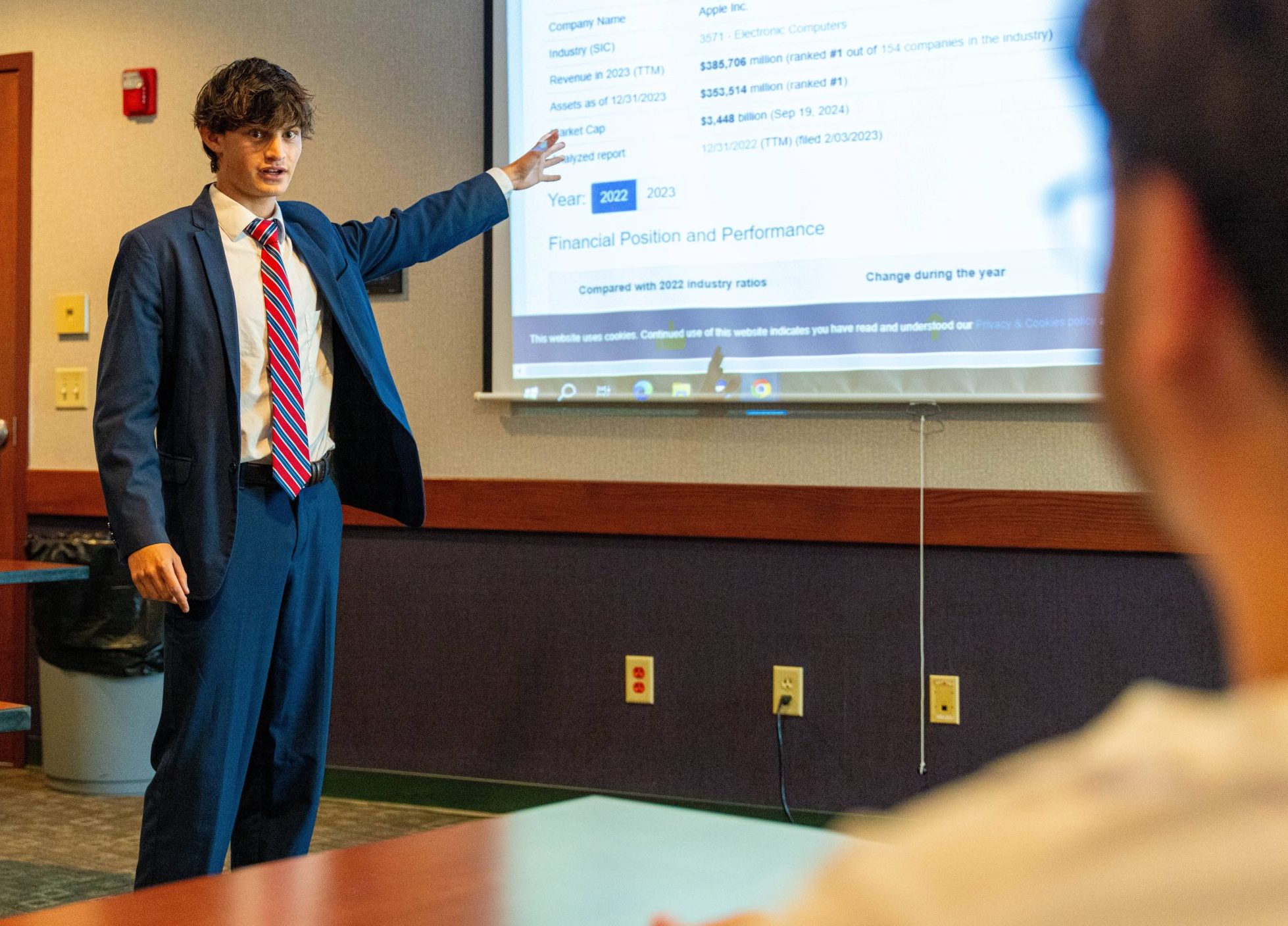 male student in suit gesturing toward image on a wall screen