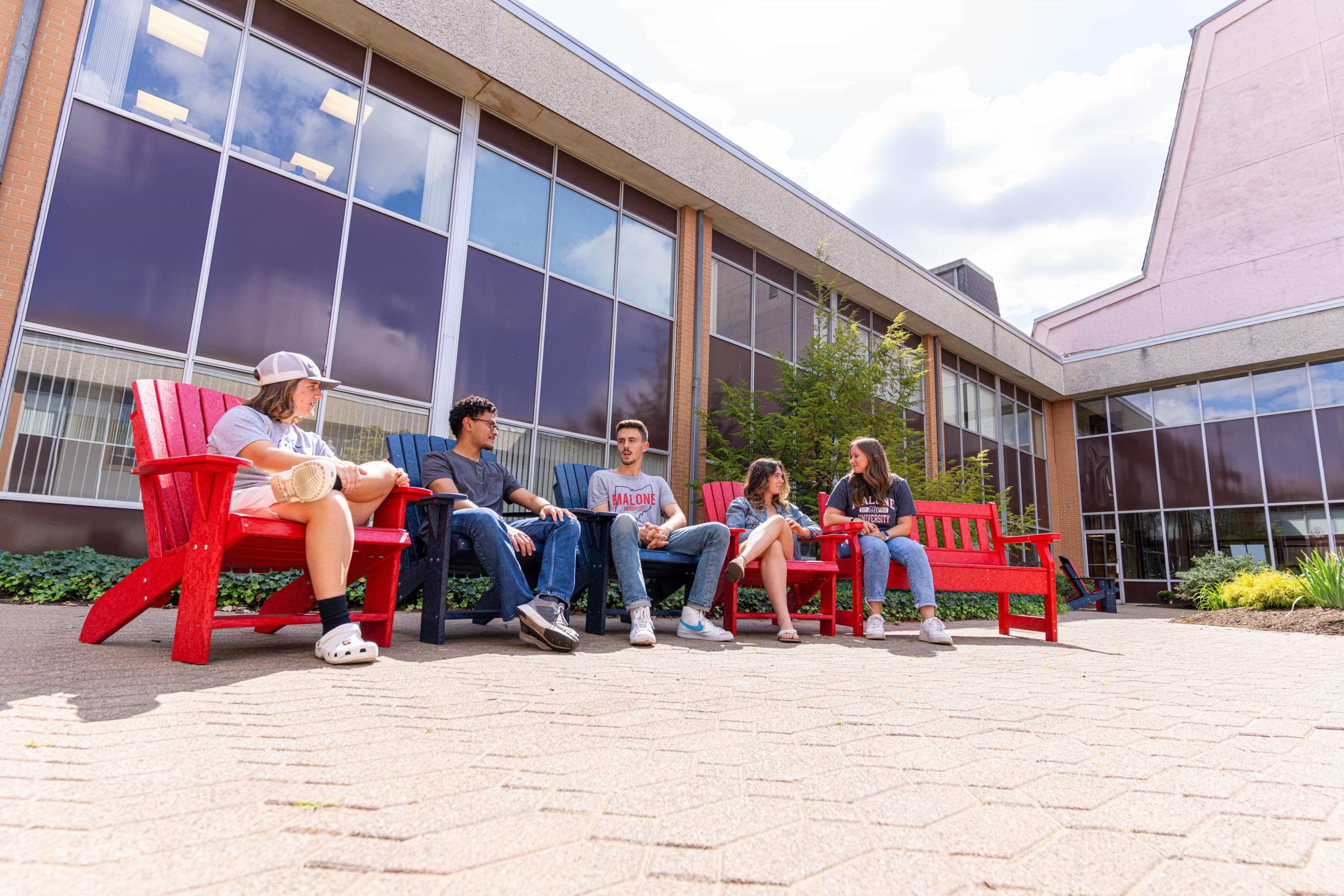 5 students outdoors sitting in red and blue chairs chatting with each other
