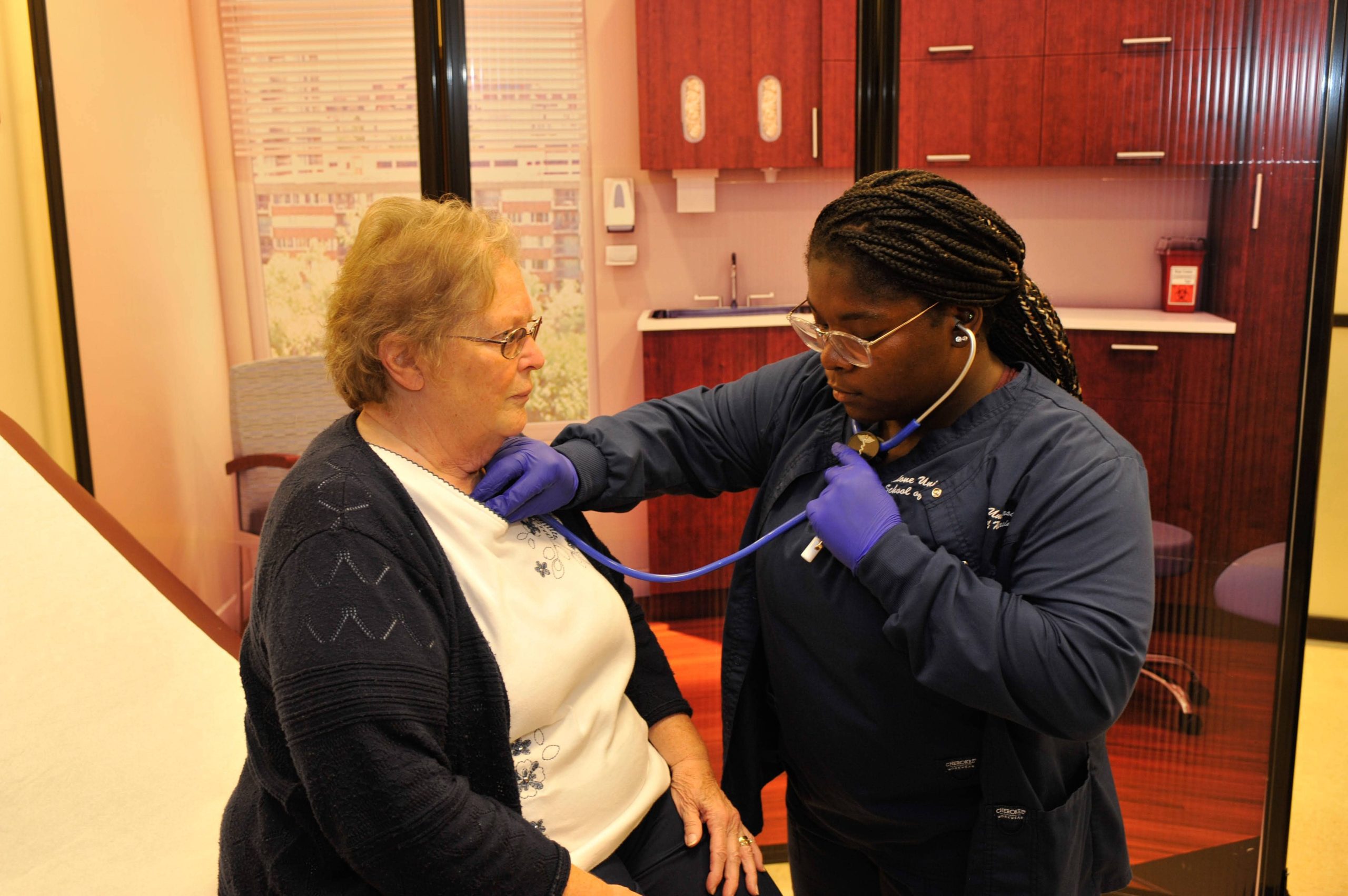 female nursing student dressed in blue scrubs listening to a patient's heartbeat