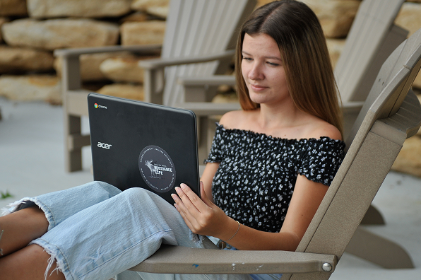 Girl in an off the shoulder top sits outdoors in a chair using a laptop