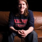 Male student sitting on a couch smiling at camera in a Malone shirt