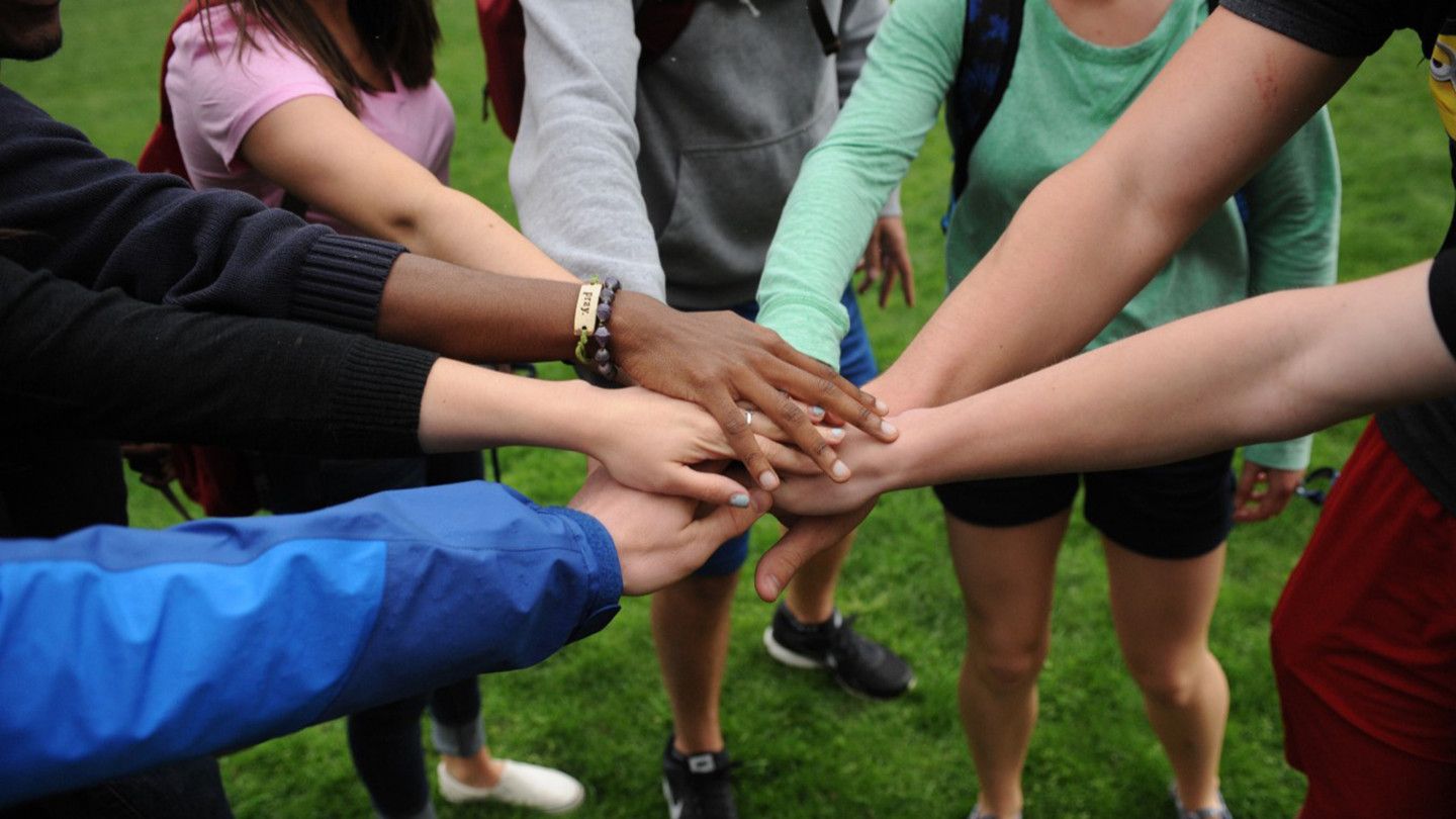 Eight students place their hands in the middle of a circle as a group