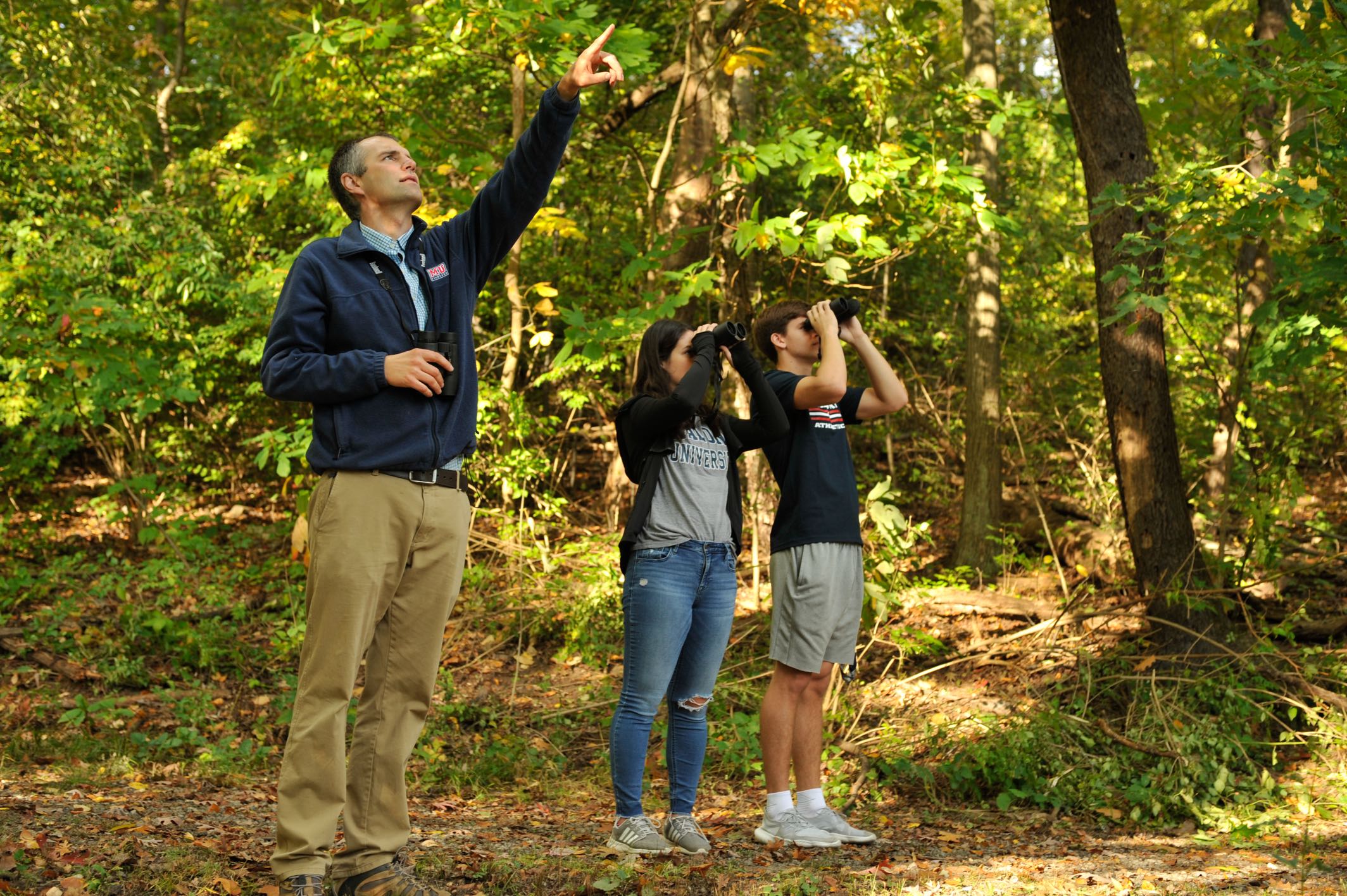 2 students and professor looking through binoculars in the woods