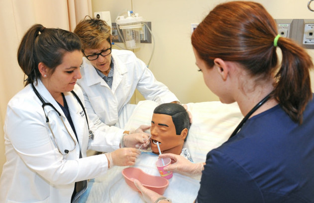 A nursing student and faculty members, wearing lab coats and scrubs, work on a mannikin in a nursing lab