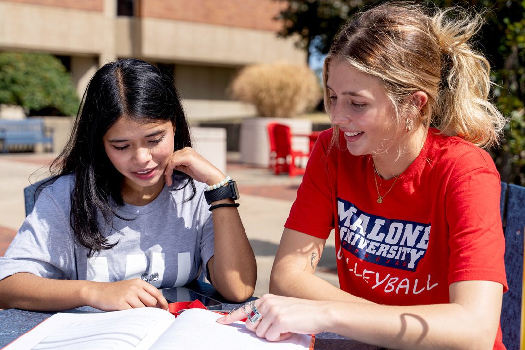 2 females viewing an open book at outdoor table in front of Cattell Library