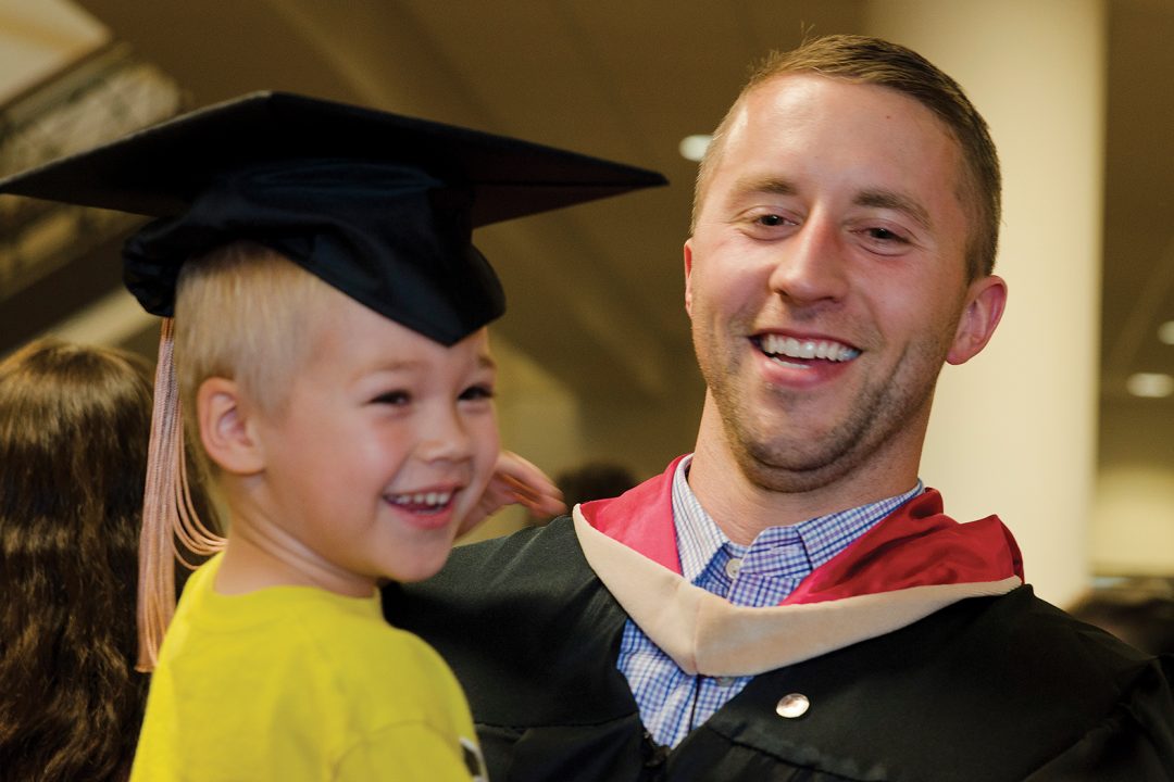 smiling male graduate student holding young child on whose head is the graduate's mortarboard