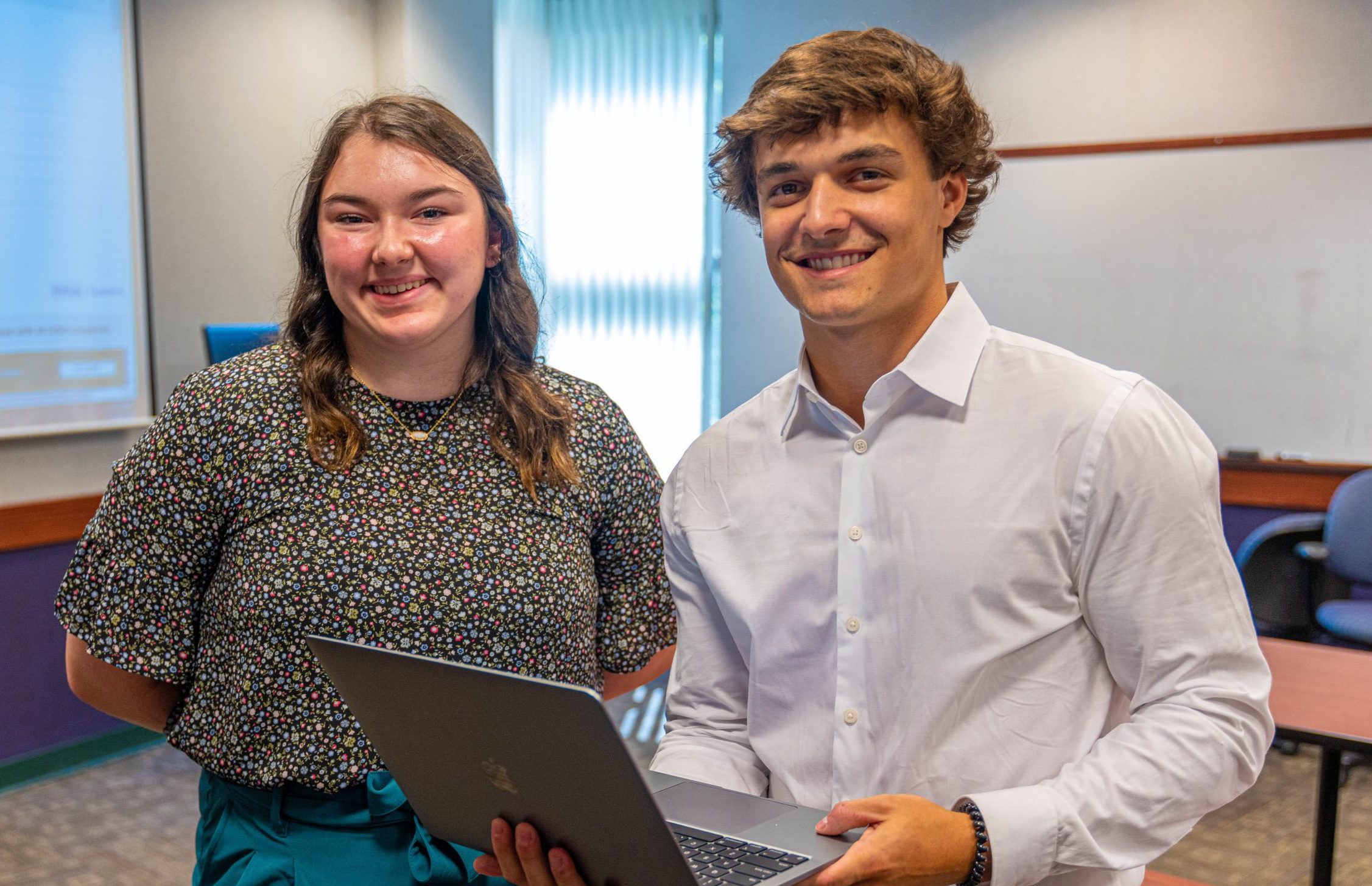 2 students standing in Mitchell 201, one of whom is holding an open laptop computer