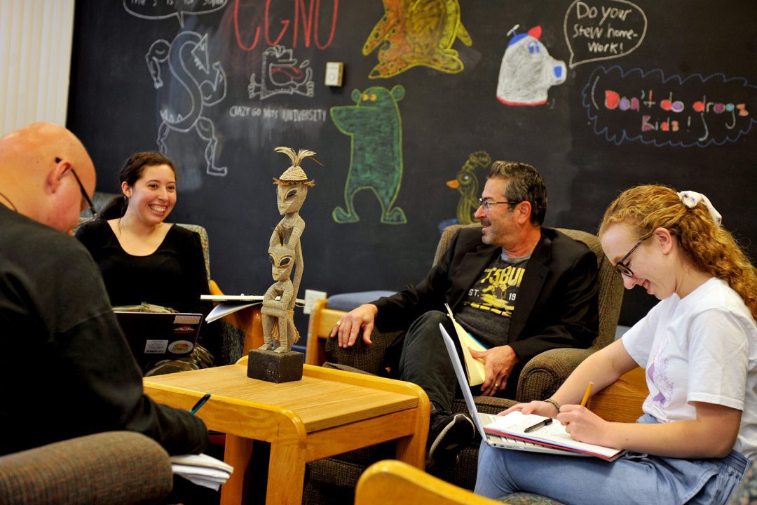 1 instructor and 3 students clustered in circle of large chairs with humorous drawings on chalkboard behind them