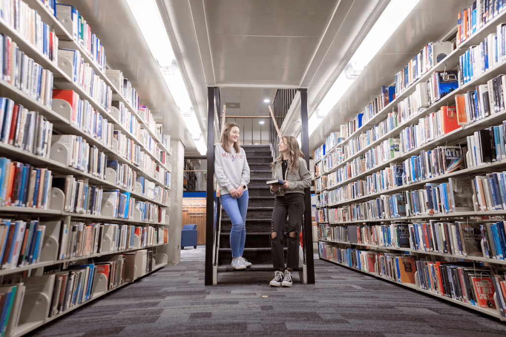 Two girls stand on the stairs in between rows of books inside the Malone library