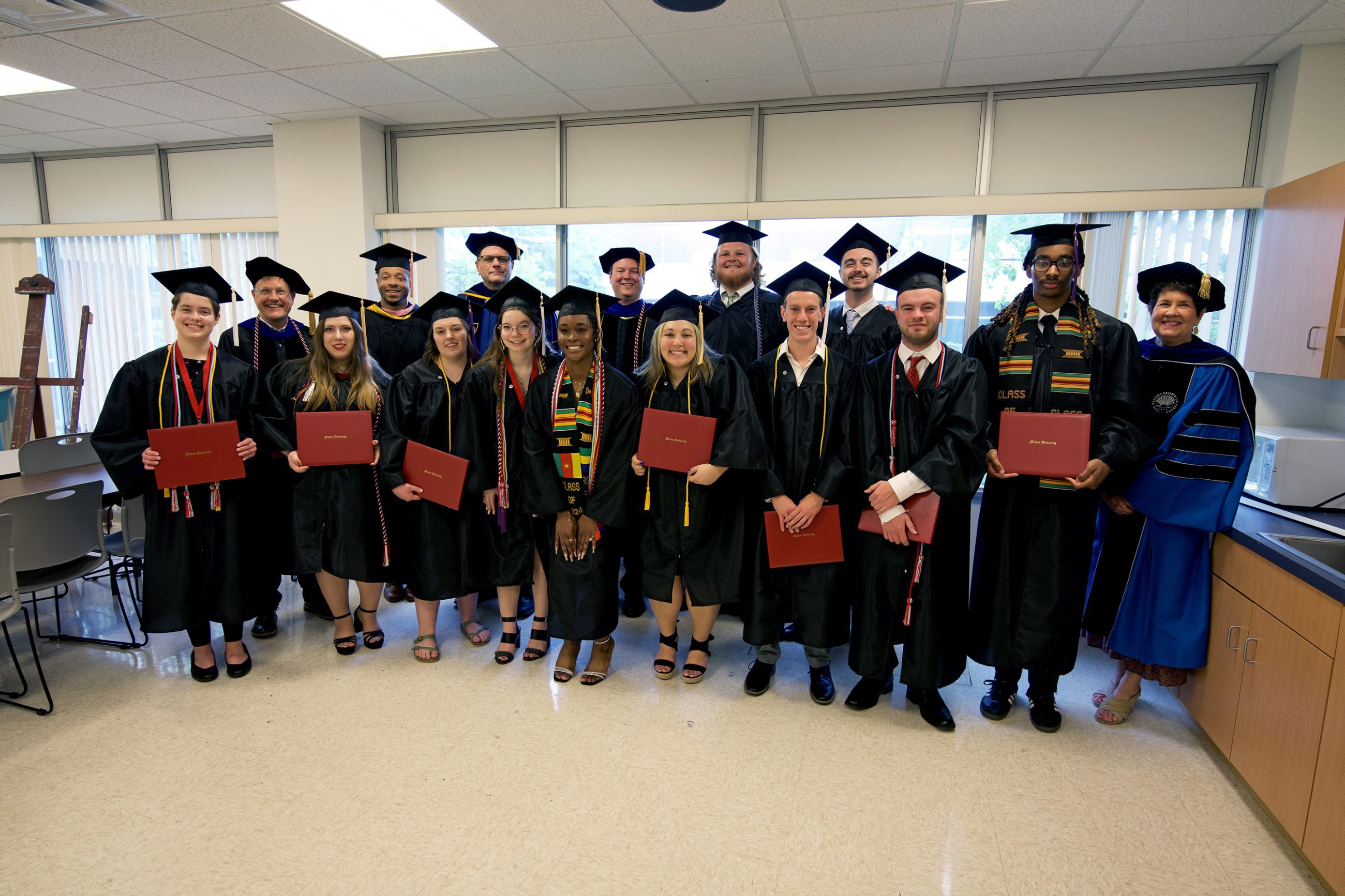 group of HEM faculty and graduates dressed in academic garb; students proudly holding diploma covers